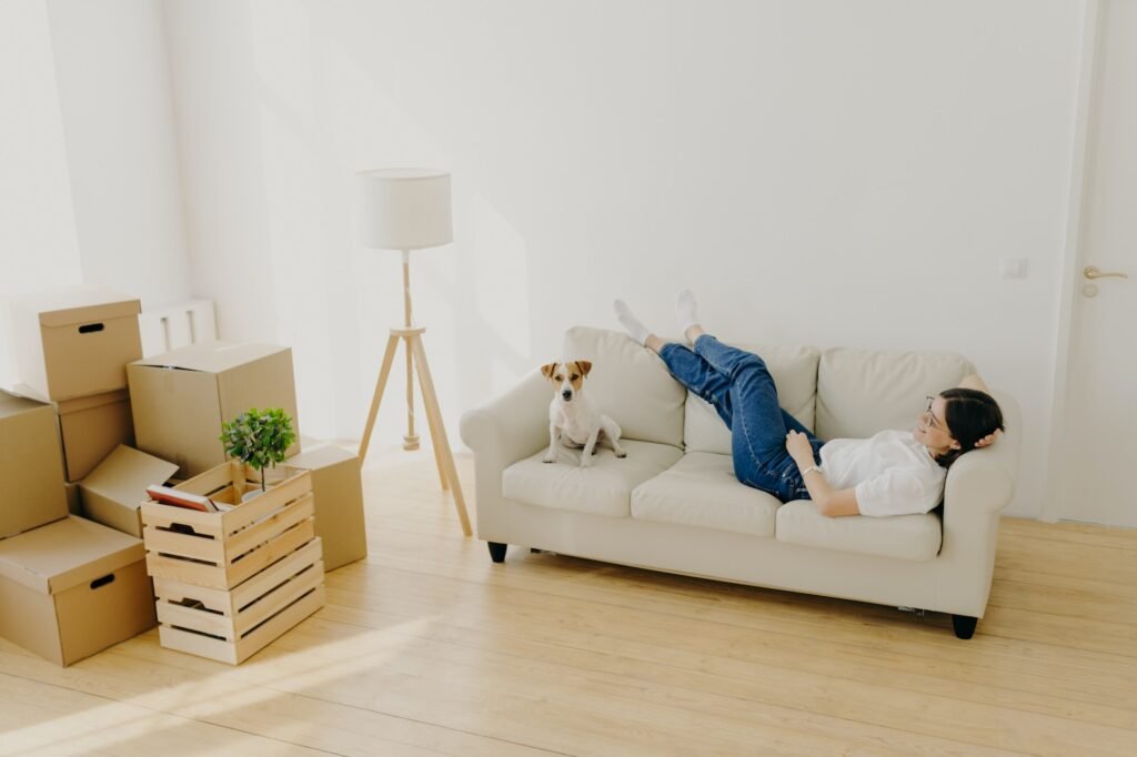 Female renter lies on white comfortable couch, raises crossed legs, poses with favourite dog